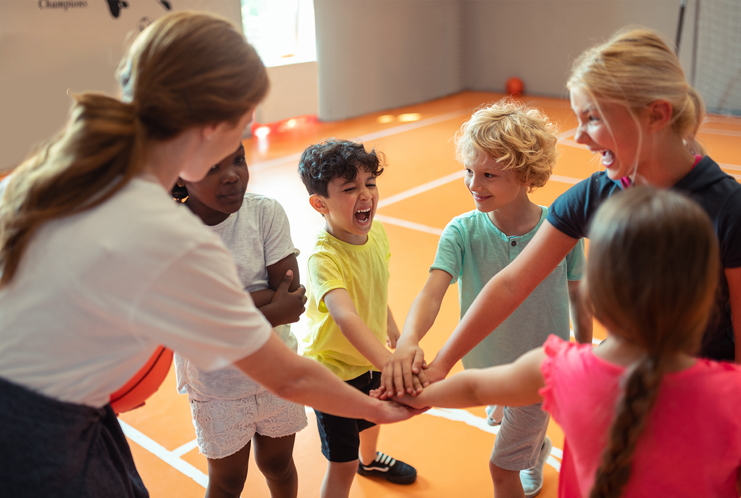 In einer Turnhalle stehen mehrere Kinder und zwei Trainerinnen im Kreis, legen in der Mitte ihre Hände aufeinander und rufen etwas
